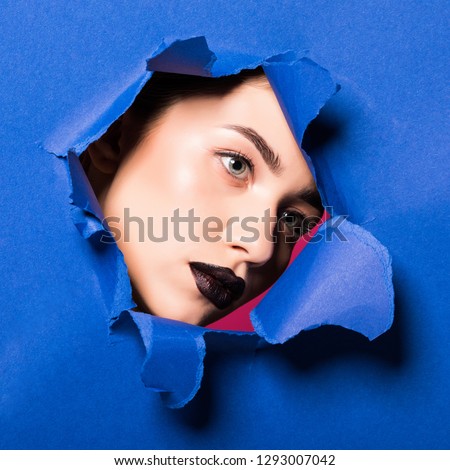 The face of young beautiful girl with a bright make-up and puffy dark lips peers into a hole in blue paper.