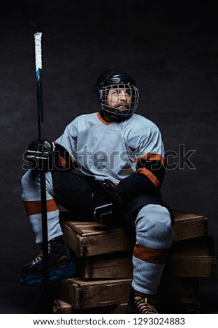 Bearded hockey player wearing full sports equipment holding a hockey stick while sitting on wooden pallets. 
