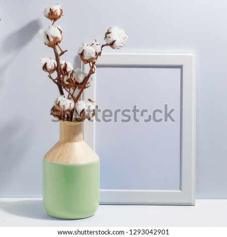 Mock up white frame and dry cotton twigs in blue ligth-green on book shelf or desk. Minimalistic concept.