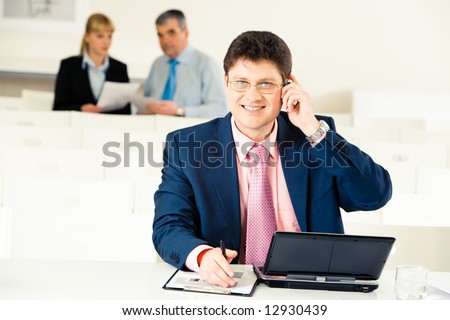 Portrait of confident man calling on his mobile phone and looking at camera with laptop and some documents near by
