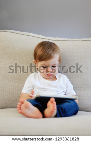 Cute little baby boy with funny face expression watching cartoons on digital tablet sitting on sofa at home. Copy space. Child and gadget, children internet safety concept. Parenting control.