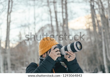 A man or guy in winter clothes and vintage attire taking pictures with his camera, DSLR and telephoto lens of the outdoor and mountain peaks. 