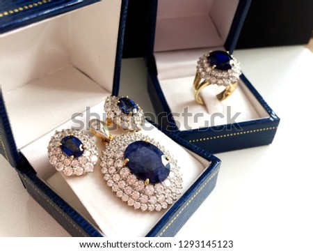 Pendant earrings and ring set of blue sapphire egg shape surrounded by diamond in blue jewel box side view on black and white background.