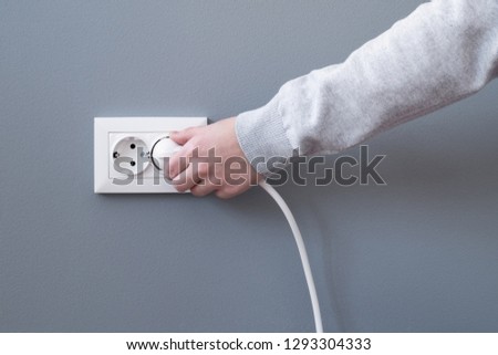 Hand plugging in an electric cord into a white plastic socket or  european wall outlet on grey plaster wall. Closeup of a woman's hand inserting an electrical plug into a wall socket. Daylight.  Royalty-Free Stock Photo #1293304333