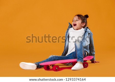 Stylish little child girl in jeans clothes sitting on skateboard on orange background