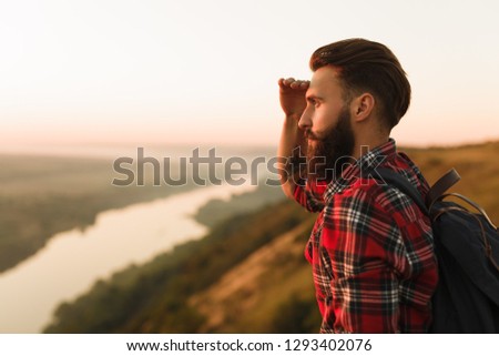 Handsome bearded guy with backpack keeping hand near forehead and looking at distance while standing on blurred background of evening countryside