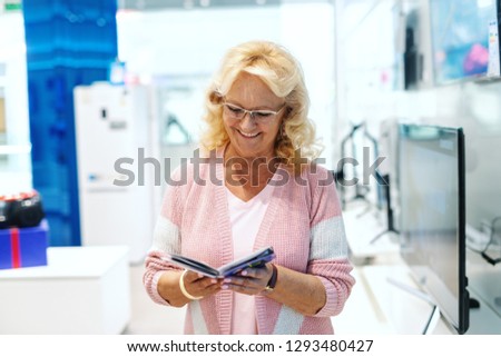 Smiling old blonde Caucasian woman with eyeglasses and dressed casual looking at brochure while standing in tech store.