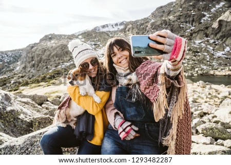 
Two young women, covered with a blanket, on a sunny winter morning enjoying a day on the mountain with their cute little dog taking themselves pictures with their smartphone. Lifestyle.