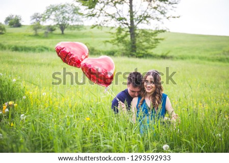 Outdoors, nature photo of young, beautiful couple holding heart shaped balloon, hiding in grass