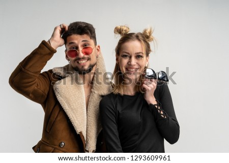 Young couple of bearded man in coat and pretty woman in black dress with long blonde hair, both with sunglasses posing in studio on gray background, copy space