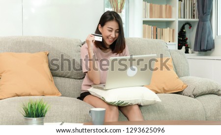 attractive young asian woman shopping online by using notebook computer and credit card, sitting on sofa in beautiful decoration living room, happy lifestyle with modern technology concept.