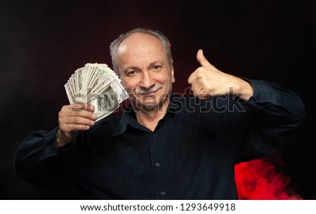 Senior gentleman holding a stack of money. Portrait of an excited old businessman. Happy old man holding dollar banknotes on dark red background. 