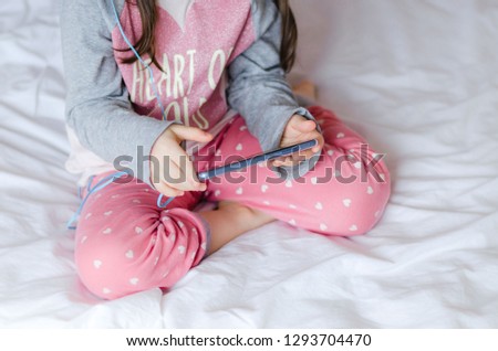 Little girl holding a smart phone in hands and sitting in bed. Little girl dressed in pink pajamas. Selective focus.Close up