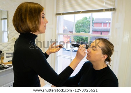 Young and beautiful professional make up artist applying make up, eyeliner and foundation to woman sitting down in chair in a private studio.