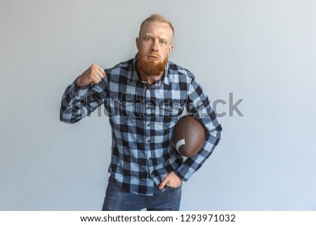 Red hair mature man standing isolated on grey wall with american football ball hand in pocket showing fist looking camera agressive