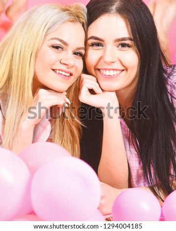 Blonde and brunette on smiling faces have fun at domestic party. Girls lay on belly near balloons, pink background. Sisters or best friends in pajamas at girlish pajamas party. Friendship concept.