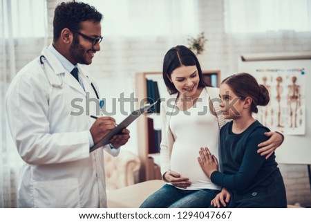 Indian doctor in white gown seeing patients in office. Pregnant woman with daughter.