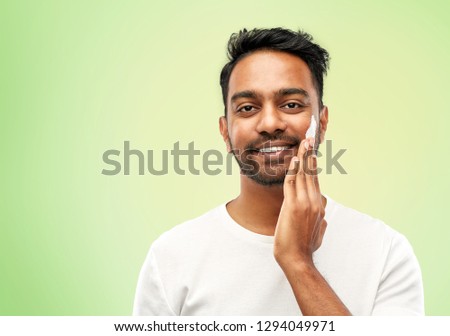 grooming, skin care and people concept - smiling young indian man applying cream to face over lime green natural background