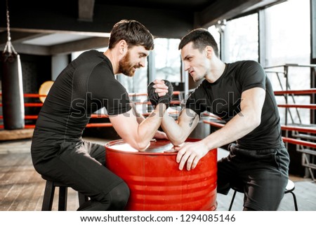 Two young athletes in black sportswear having a hard arm wrestling competition on a red barrel in the gym