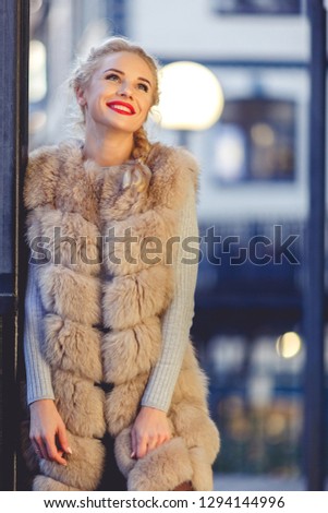 Picture of young fashionable blonde in fur vest on walk in city in evening.