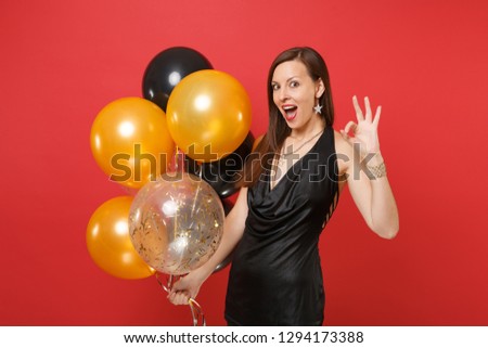 Amazed young girl in little black dress celebrating, showing OK sign, holding air balloons isolated on red background. International Women's Day, Happy New Year, birthday mockup holiday party concept