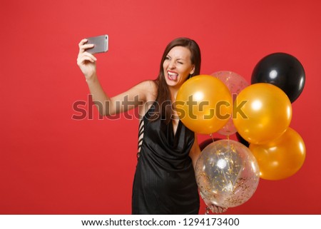 Funny young woman in black dress blinking showing tongue holding air balloons doing taking selfie shot on mobile phone isolated on red background. Happy New Year birthday mockup holiday party concept