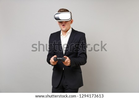Amazed young business man in classic black suit, shirt looking in headset, playing video game with joystick isolated on grey background. Achievement career wealth business concept. Mock up copy space