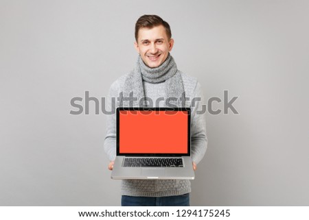 Smiling young man in gray sweater, scarf holding laptop pc computer with blank empty screen isolated on grey background. Health, online treatment consulting, cold season concept. Mock up copy space