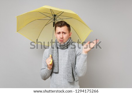 Upset young man in gray sweater, scarf holding yellow umbrella, spreading hand isolated on grey background. Healthy fashion lifestyle, people sincere emotions, cold season concept. Mock up copy space
