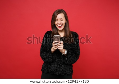 Laughing cute young woman in black fur sweater using mobile phone, typing sms message isolated on bright red wall background in studio. People sincere emotions, lifestyle concept. Mock up copy space