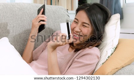 Asian attractive young woman shopping online by using tablet and credit card, sitting on light brown sofa in beautiful decoration living room, happy lifestyle with modern technology concept.