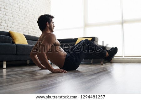 Shirtless fitness instructor performs a knee tuck exercise at home. Muscular young good looking white male is working out on the floor of his house doing an ABS routine and training his core muscles. Royalty-Free Stock Photo #1294481257