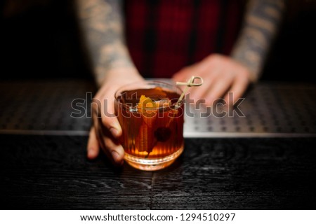 Bartender serving a glass of a Old Fashioned cocktail with orange zest on the bar counter on the blurred background Royalty-Free Stock Photo #1294510297
