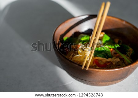 Chicken Stir Fry with Rice Noodles in a wooden bowl and chopsticks on a stone background with sunny stunning long shadows with copy space. Asian cuisine.