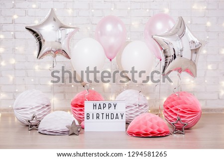 birthday party concept - close up of air balloons, paper balls and lightbox with happy birtday text over brick wall with lights
