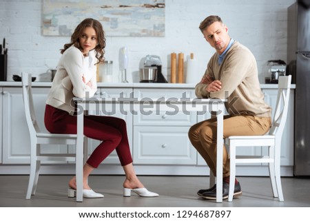 dissatisfied couple sitting with arms crossed at table in kitchen