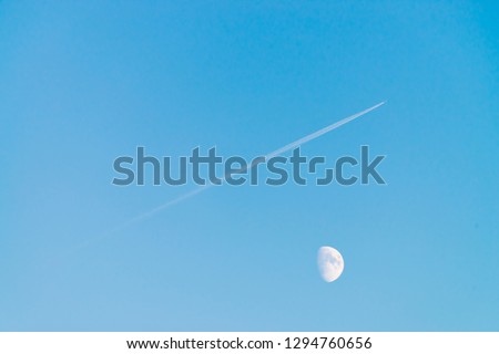 Condensation track of jet above moon in clear blue day sky. Minimalist blue background. Plane flies up diagonally. Airplane is flying in air space. White moon in cyan sky.