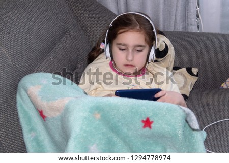 girl in headphones playing in the phone, lying on the sofa, the concept of childish play addiction, natural photo