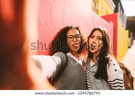 Happy young girls having fun outdoors and taking selfie. Excited female friends showing their coloured tongue after eating a candy.