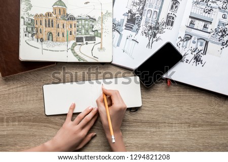 top view of womans hands drawing on paper, albums with paints and smartphone on wooden background