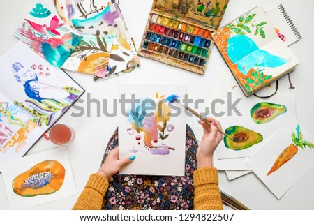 top view of woman holding drawing on knees and painting in it with watercolors paints while surrounded by colored pictures