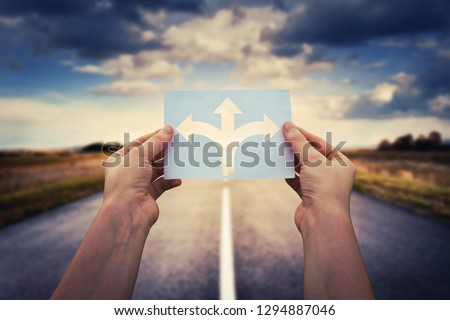 Hands holding paper with arrows crossroad symbol splitted in three different directions. Choose the correct way between left, right and front. Difficult decision concept, over asphalt road background. Royalty-Free Stock Photo #1294887046