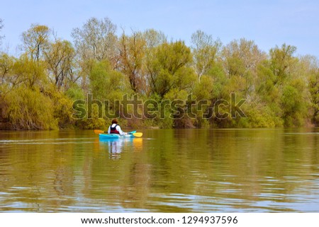 Rear view of woman paddle blue kayak on Danube river against the background of light green spring tree. Spring kayaking. Concept for adventure, travel, action, lifestyle.