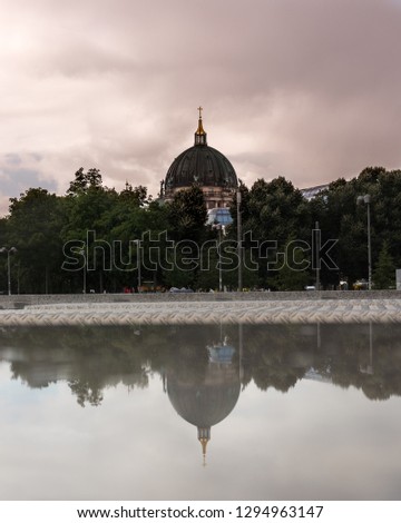 This is a photo of a famous church in Berlin with a big cupola hiding behind the trees in the picture. It is raining and cloudy and there's a reflection of the church at the bottom of the picture.