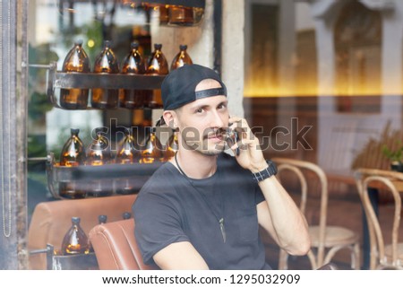 Picture through glass window of handsome glad Caucasian man with beard and mustache,smiling gently at camera  while talking on mobile phone, having playful look, listening to interlocutor attentively