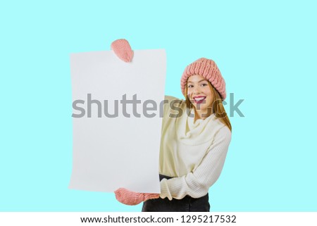 A girl with a poster in her hands. A young red-haired girl in a knitted pink hat is holding a poster with both her hands on the side of herself and smiling looking at the camera against an isolated
