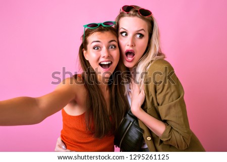 Two happy pretty hipster girls making selfie with funny surprised emotions, natural make up, vintage outfits, studio pink background.