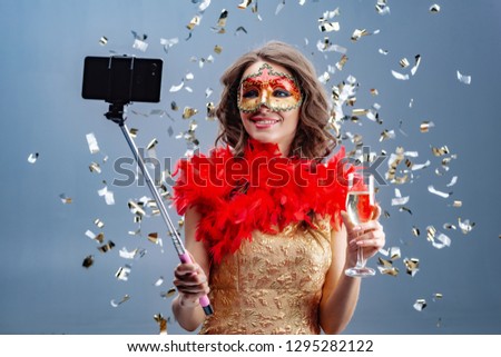 Photo of a smiling woman in  carnival mask and  gold dress with  raised glass makes  selfie on a mobile phone. Background with tinsel
