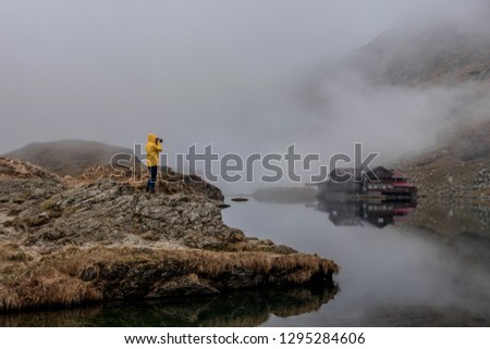 Female travelling photographer in a yellow jacket taking pictures and standing on a rocky cliff near the Balea  glacier lake in Fagaras mountains, Romania