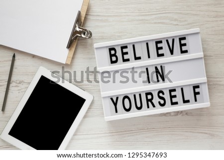 Modern board with text 'Believe in yourself', tablet, clipboard and sheet of paper on a white wooden surface, top view. Overhead.